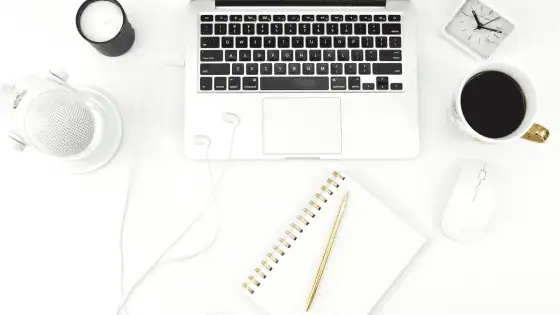 White desk with microphone, hot drink, notepad, clock, candle, and laptop.