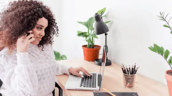 A woman is sitting at a desk in front of her computer and is talking on the phone. There are some indoor pot plants for decoration.