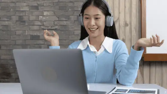 Asian woman smiling wearing headset and sat teaching online in front of a laptop
