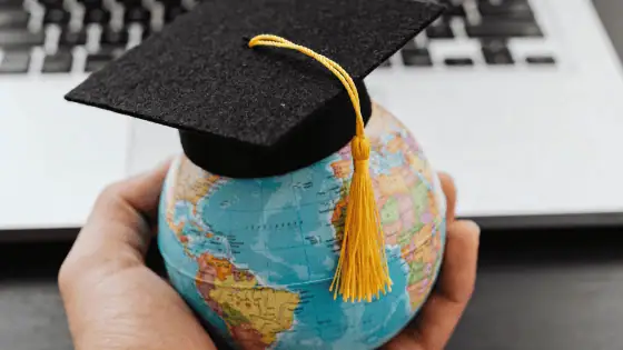 open laptop with a hand in front of it holding a small globe wearing a degree hat
