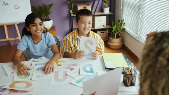 two children smiling with holding paper with letters from the English alphabet. The desk in front of them has more letters of the English alphabet for them to choose.