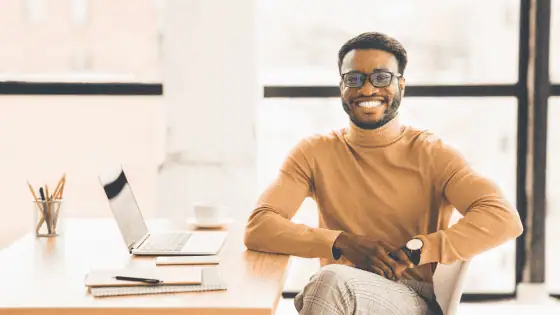 A confident black man smiling at the camera while sat at his desk.