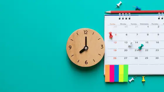 A flat lay image with a blue background showing a wooden clock next to a wall calendar and sticky notes.