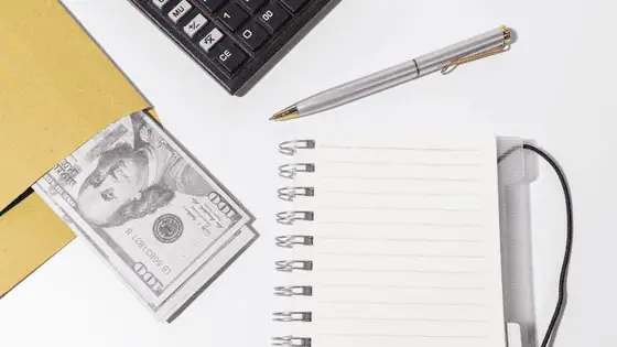 A flat lay image of a desk with money, a pen, notebook, and calculator